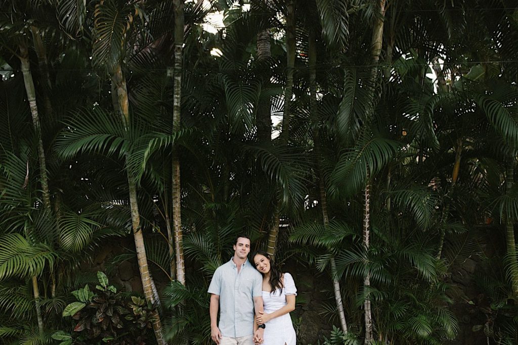 A couple stands holding hands smiling in front of a palm trees during their engagement session in Hawaii.