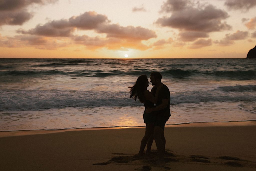 An image of an engaged couples' silhouette's embracing on a Hawaii beach for their engagement session.  There are large waves in the background.