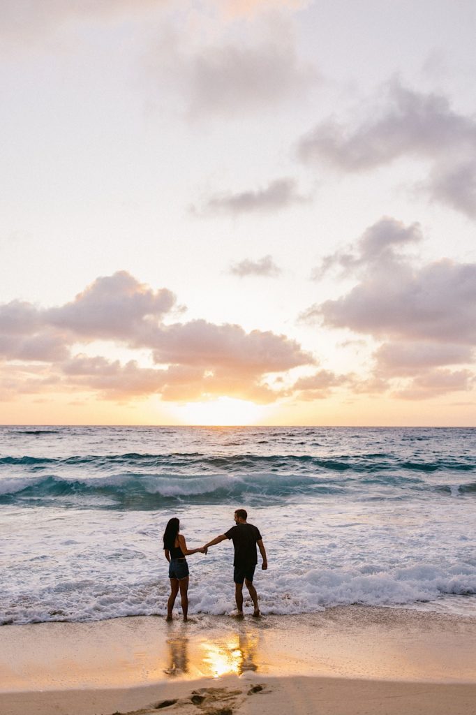 A couple walking in towards the water during their beach engagement session.  The sky is colorful as the sun begins to set.  