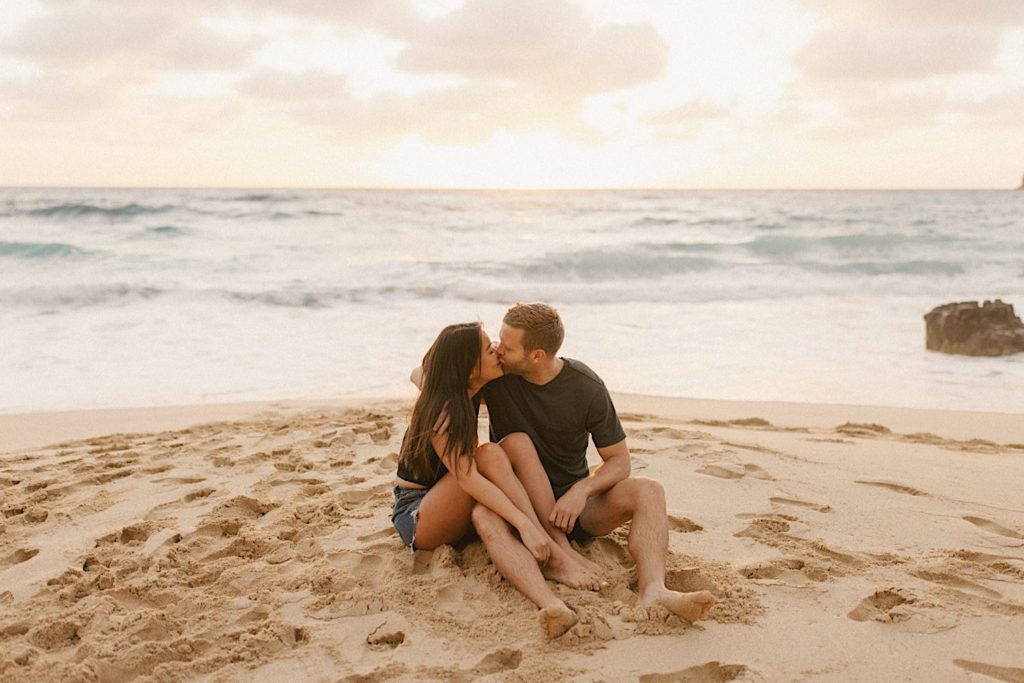 A couple sits in the sand with their legs overlapping while kissing.  The tide is bring the water up the shore behind them.