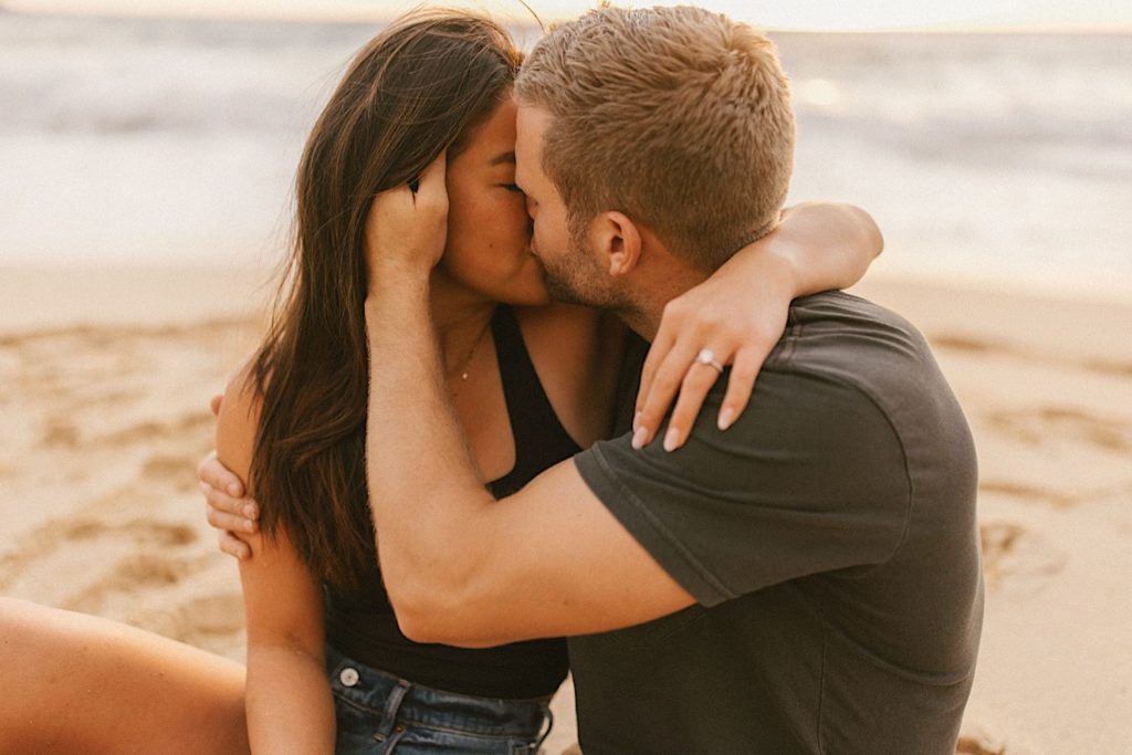 A couple kisses while holding one another sitting in the sand during their engagement session on an Oahu beach.