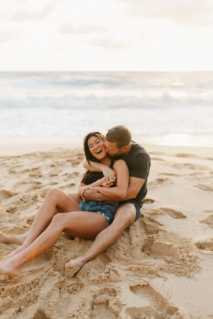 A fiancé kisses his fiancées' neck while the hold one another on a Hawaii beach for their engagement session.