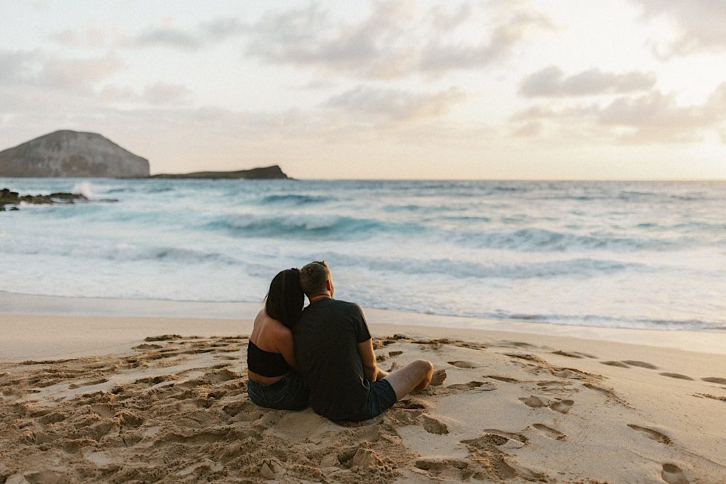 An engaged couple sits together leaning against each other while watching sunset on the coast of Oahu.
