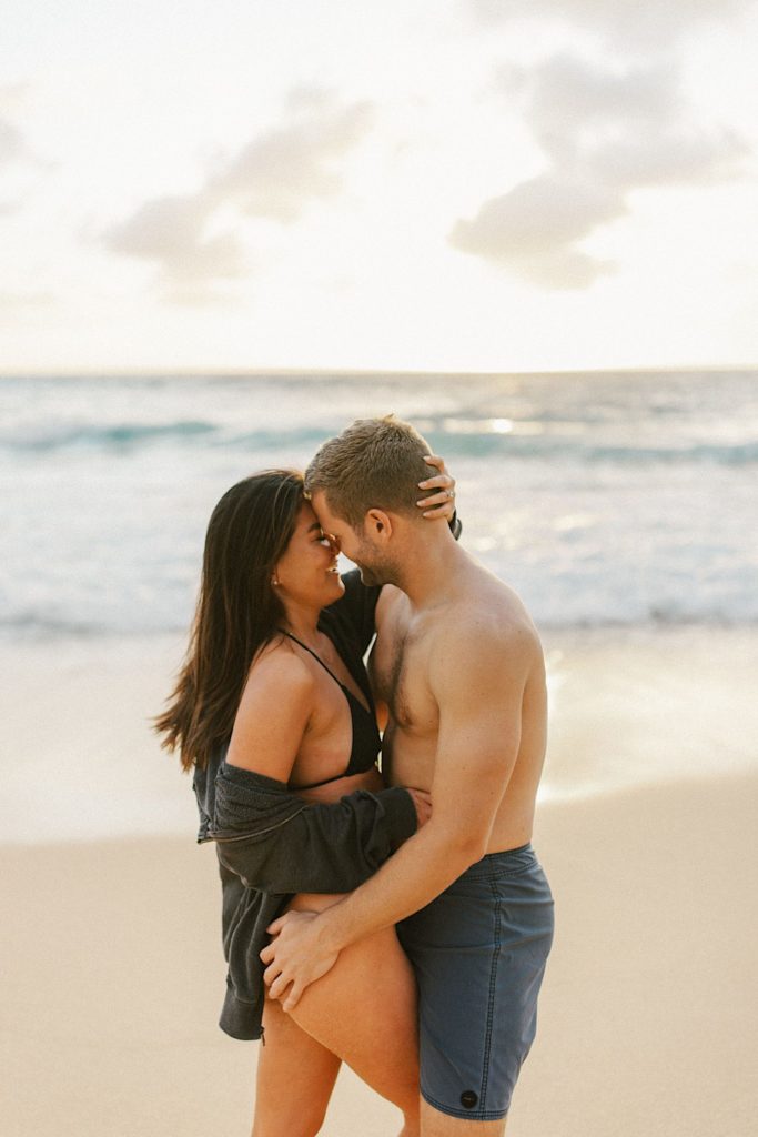 A couple poses in their swimsuits, embracing during their engagement session on a beach on Oahu.