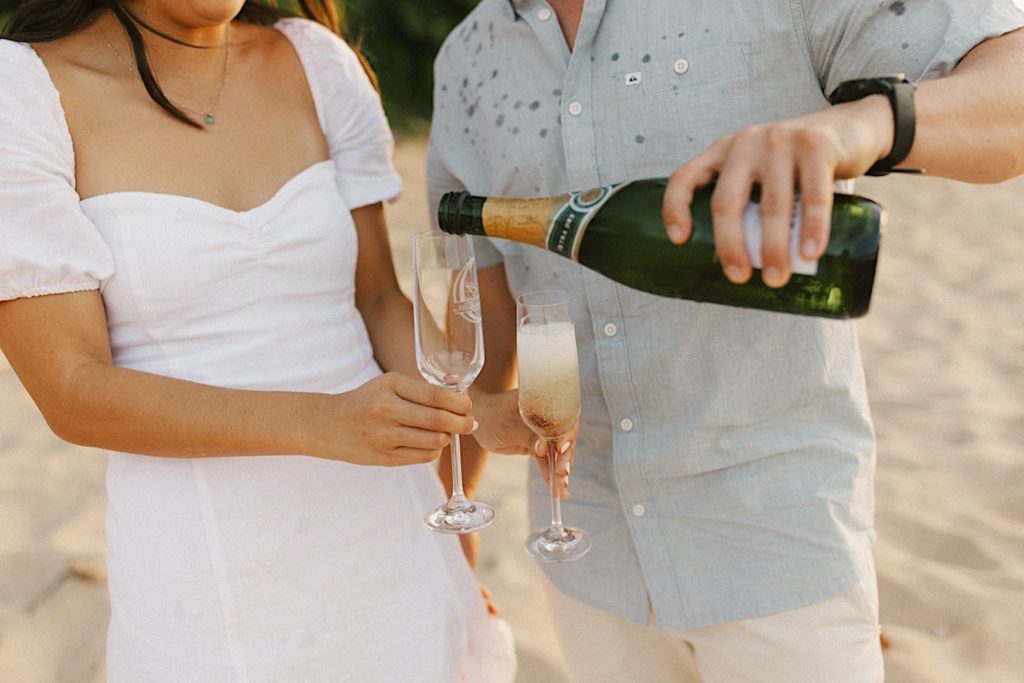 An engaged couple holds custom champagne glasses while pouring champagne on a beach.  