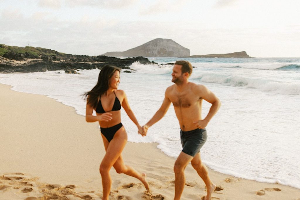 A couple runs on an Oahu beach while looking at one another during their engagement session.