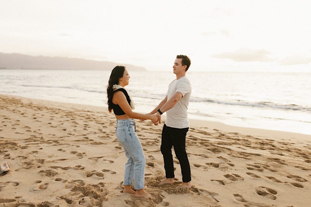 A fiancée folds the hands of her fiancé on Oahu beach during golden hour.