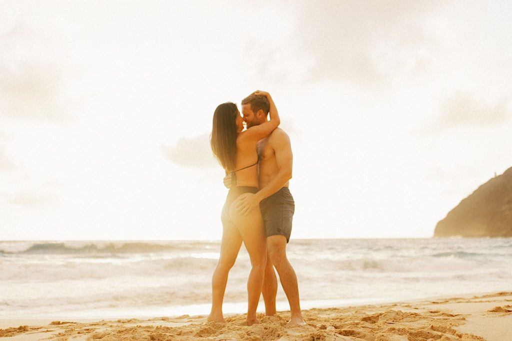 A couple holds one another while standing on a beach in Oahu during golden hour.