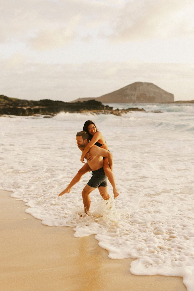 A fiancée rides on her fiancé's back during their engagement session on an Oahu beach.