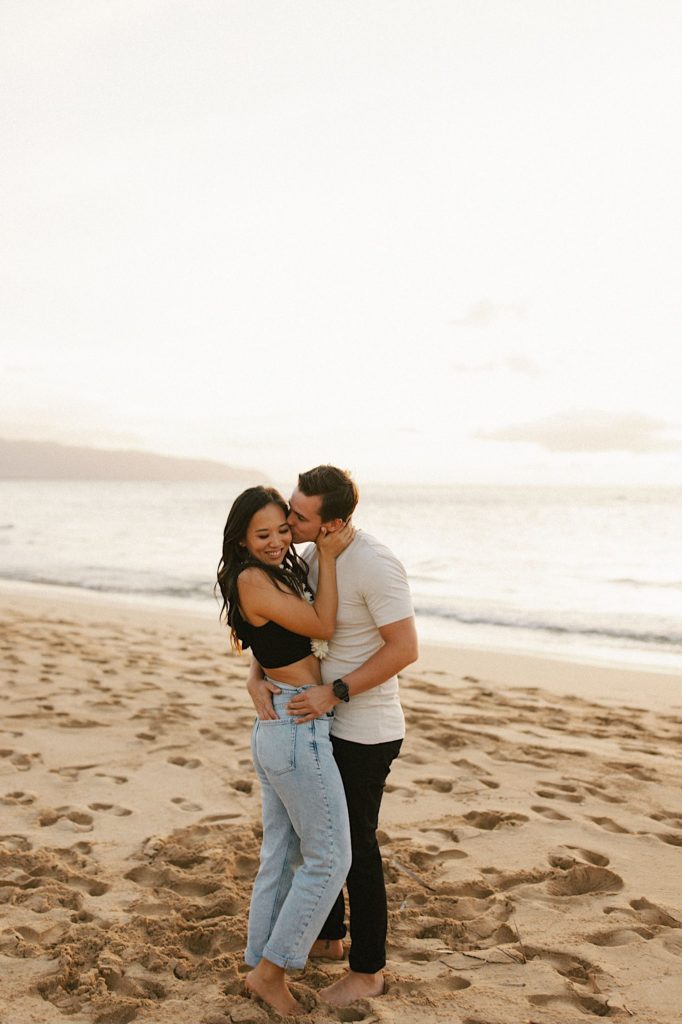 A fiancé kisses the cheek of his fiancée while they stand on a beach during golden hour in Hawaii