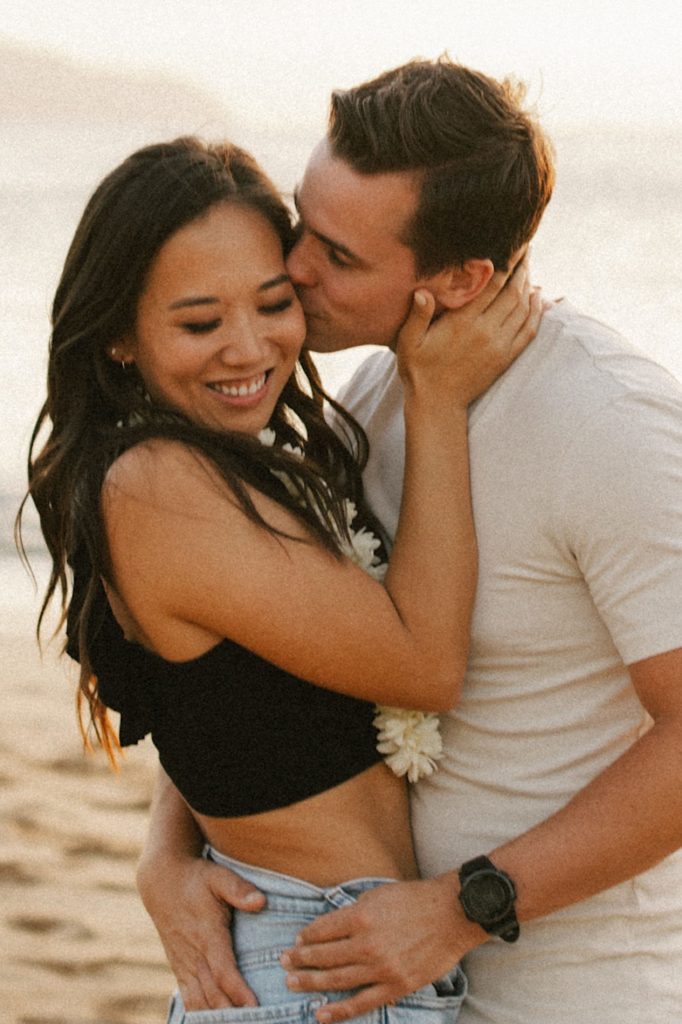 A fiancé kisses the cheek of his fiancée while they stand on a beach during golden hour on Oahu