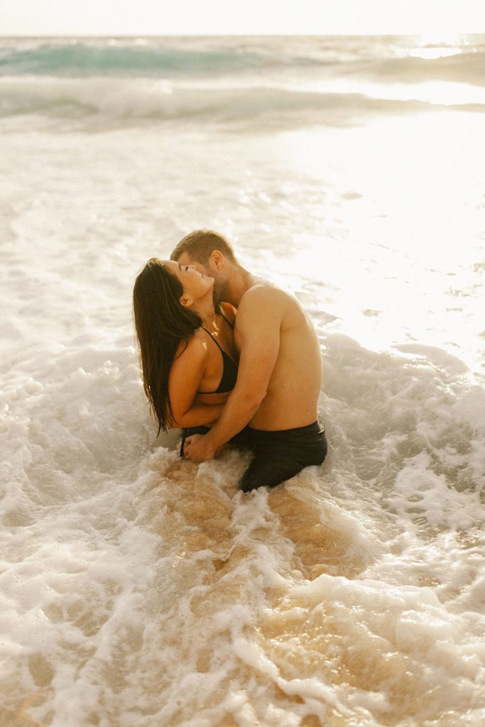 A couple holds one another and hugging and laughing in the shallow water of a beach in Hawaii