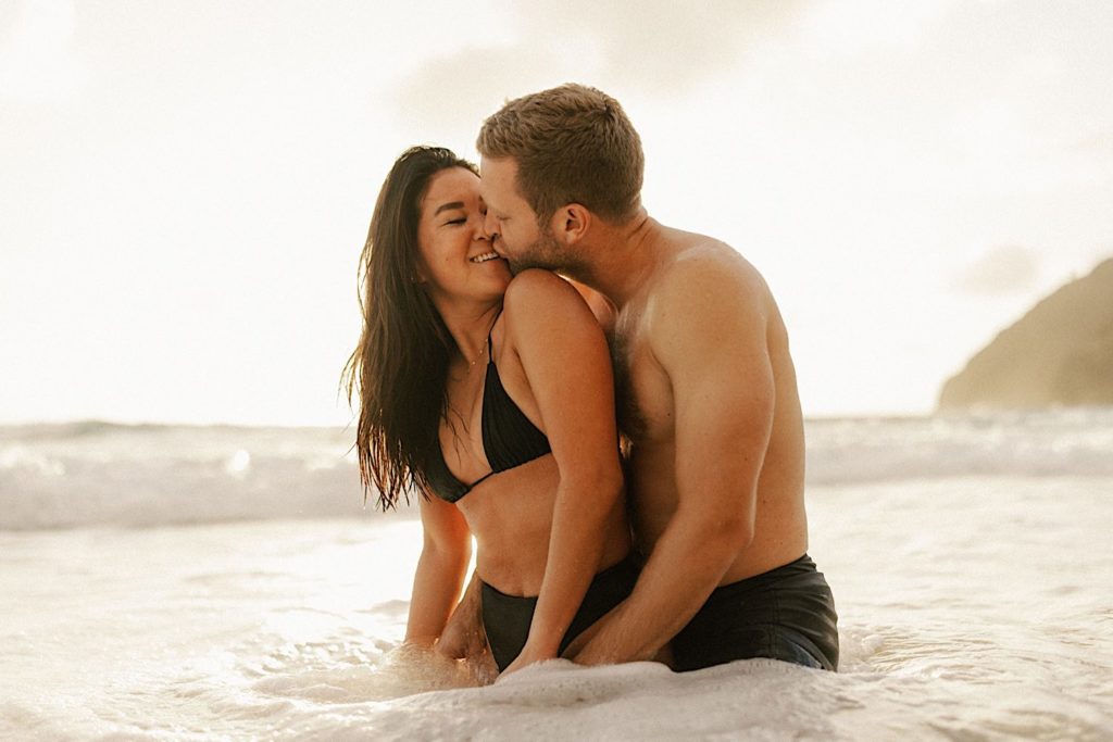 A couple holds one another kissing while sitting in the shallow waters on a beach in Hawaii.
