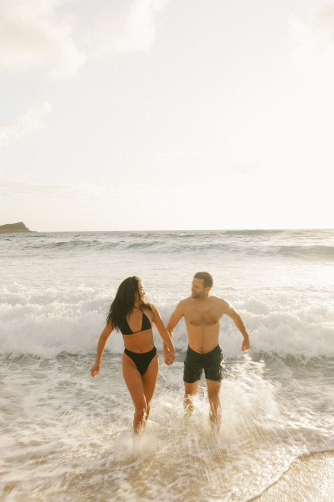 A couple walks in the water on a beach on Oahu holding hands and smiling at one another during golden hour.  