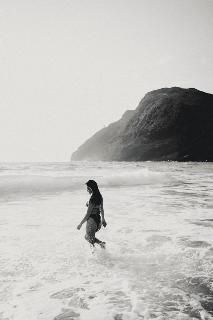 A girl walks into the water with a rocky shore in the distance on a beach in Hawaii