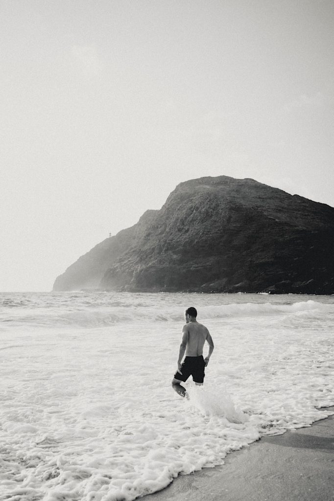 A boy walks into the water with a rocky shore in the distance on a beach in Hawaii