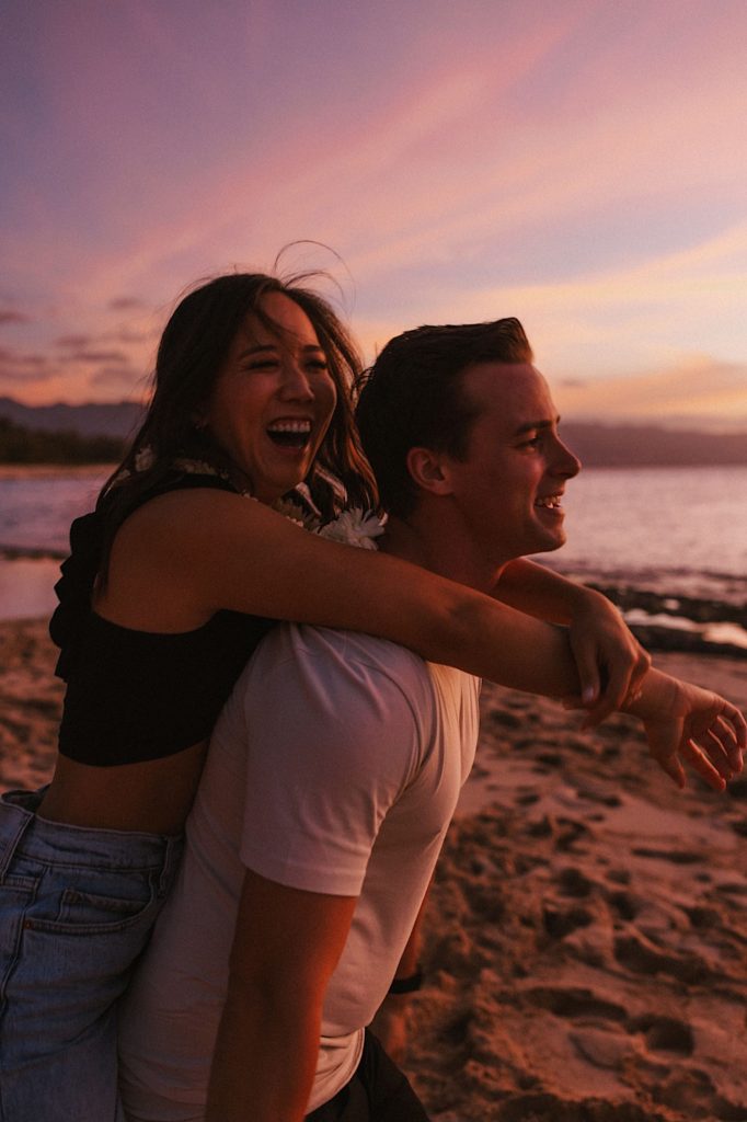 A fiancé gives his fiancée a piggy back ride while laughing during blue hour on the coast of Oahu.