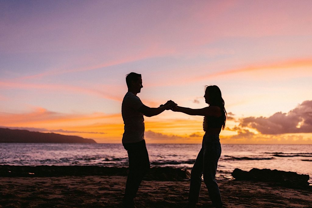 An engaged couple holds hands while dancing during sunset on a beach in Oahu.