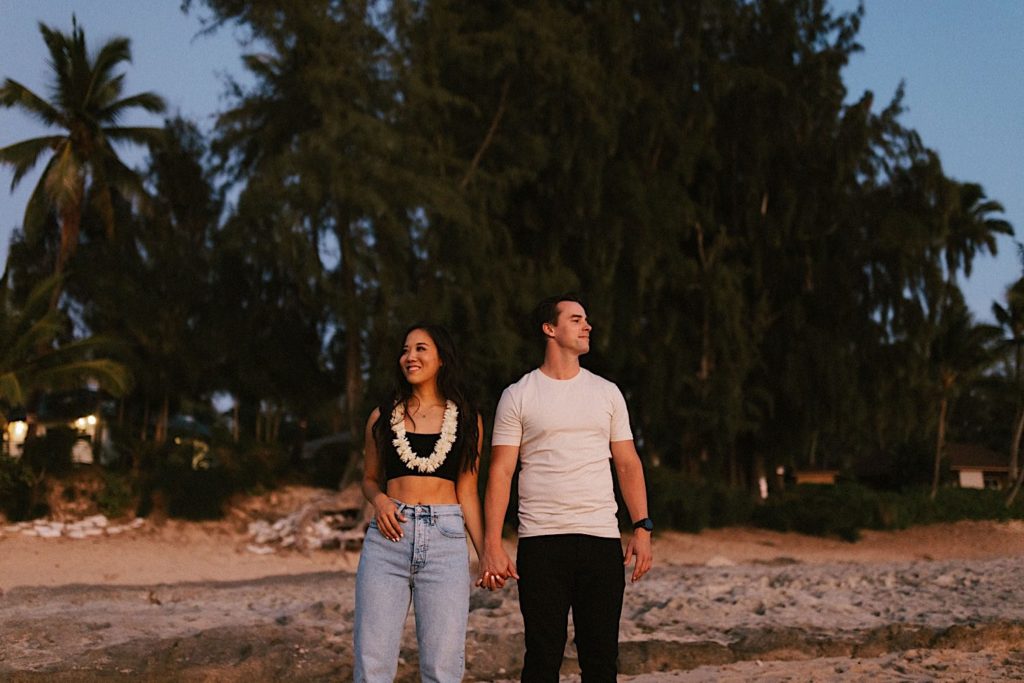 A couple holds hands and looks away from one another during blue hour during their engagement session in Hawaii.