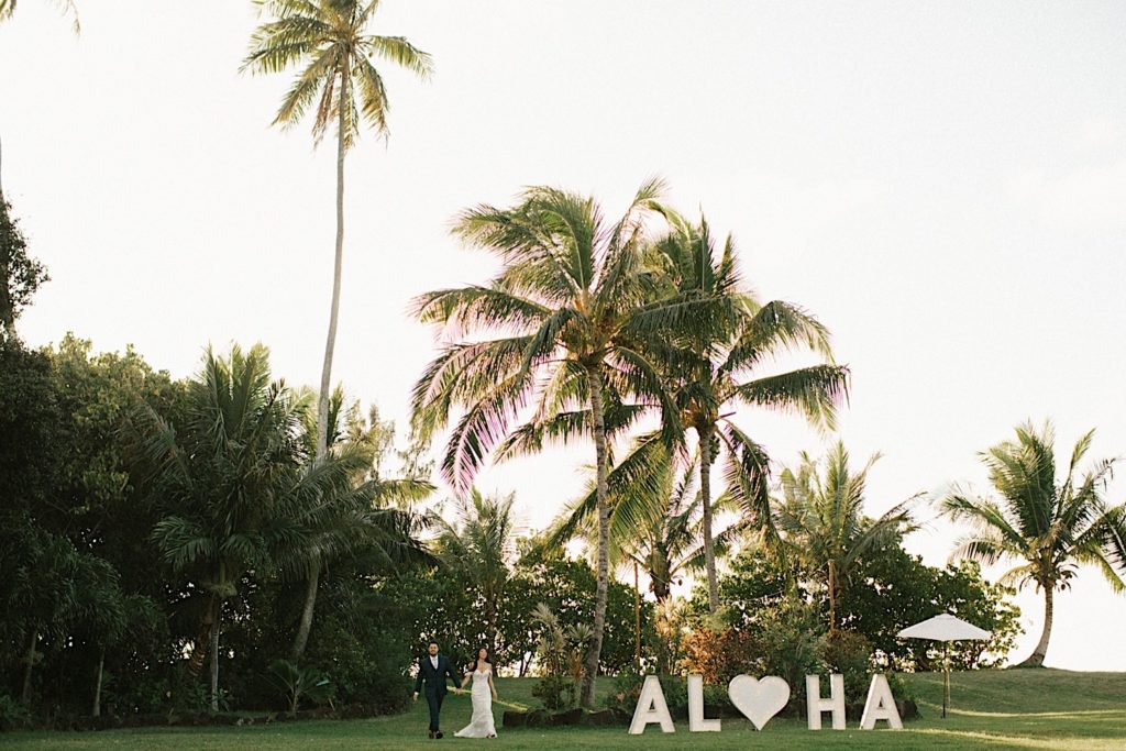 A couple walks into their wedding day next to a giant Aloha sign during their wedding at Loulu Palm in Hawaii