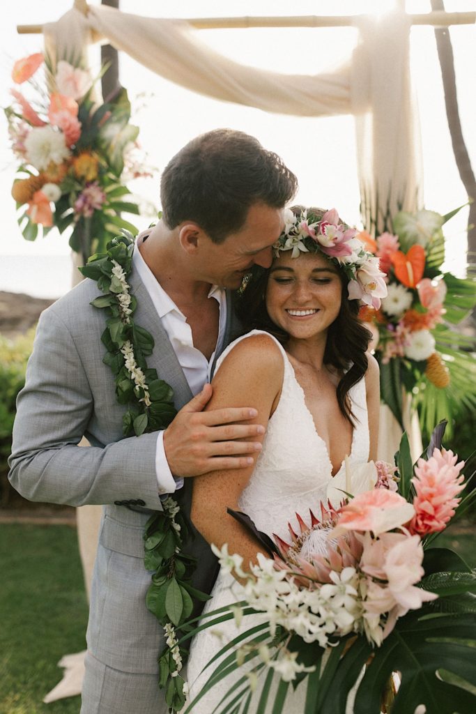 Newlyweds stand in front of their wedding arch with tropical florals draped around it.  The bride wears a tropical themed flower crown and the groom wears a traditional Hawaiian bay leaf lei
