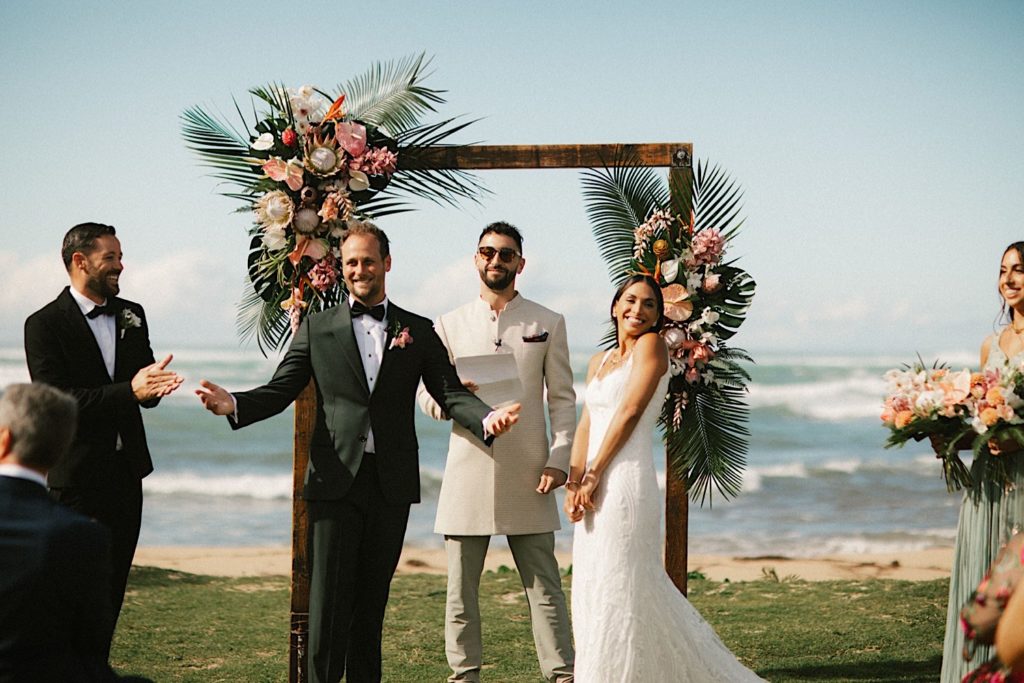 A couple smiles at their guests during their wedding ceremony standing in front of a tropical styled wedding arch.