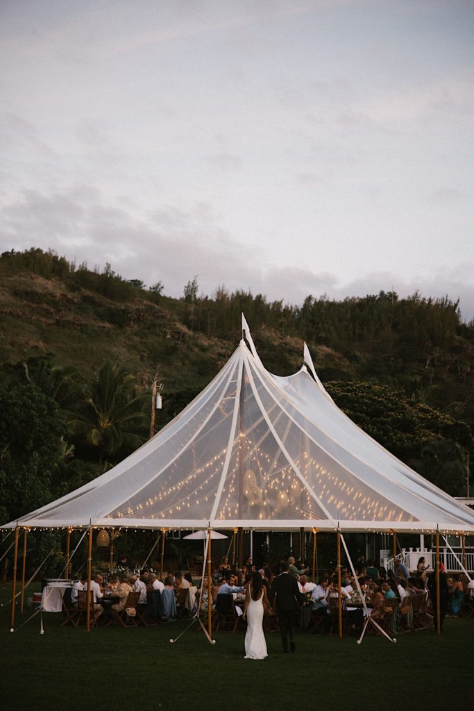 String lights hang in a clear tent as a newly married couple walks to their wedding reception at Loulu palms in Hawaii.
