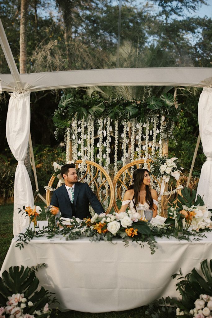 A couple sits at the head table during their wedding toasts at their tropical themed wedding in Loulu Palms in Hawaii.
