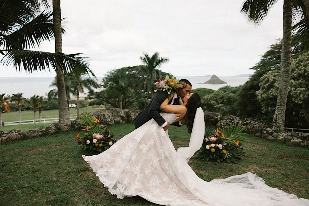 A husband kisses and dips his wife on  their wedding day while standing in front of their tropical wedding decorations.