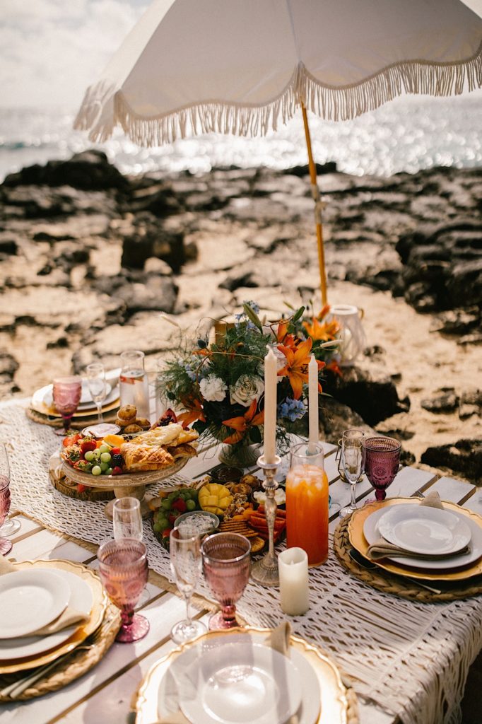 A picnic on the beach with vintage glass, a macrame table runner and a parasol with fringe to celebrate a newlywed couple.