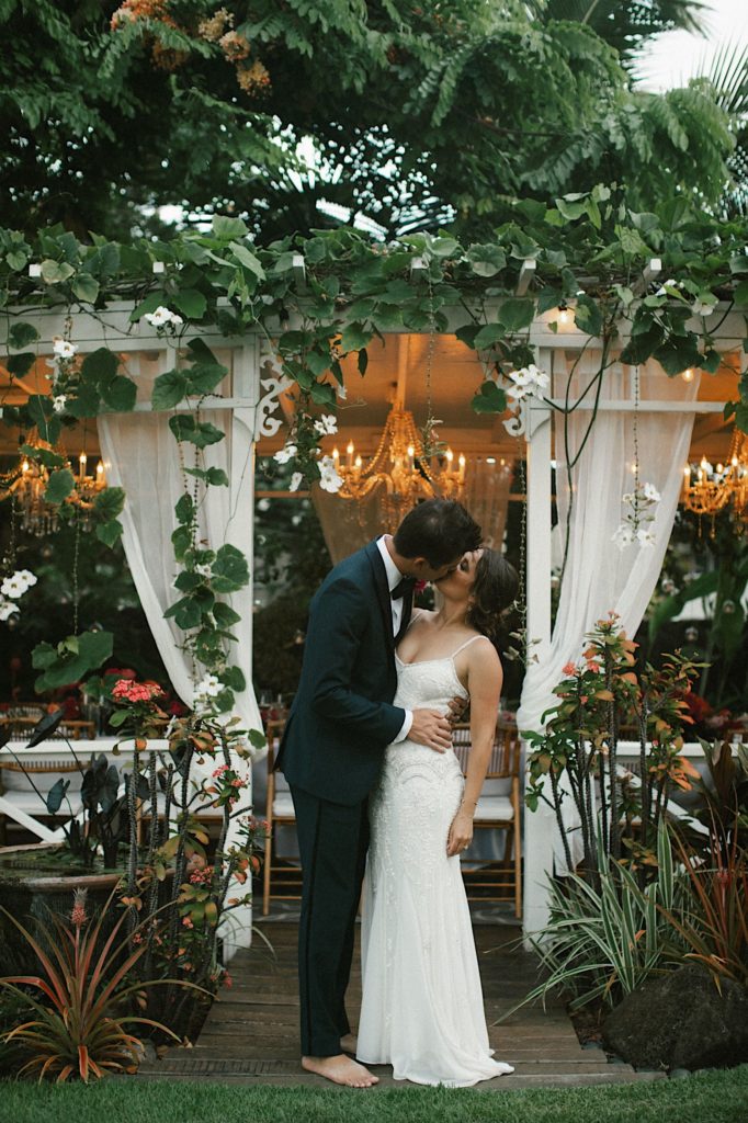 A couple stands holding one another,  and kissing in front of their reception space at the Hawaii wedding venue at Male’ana Gardens.