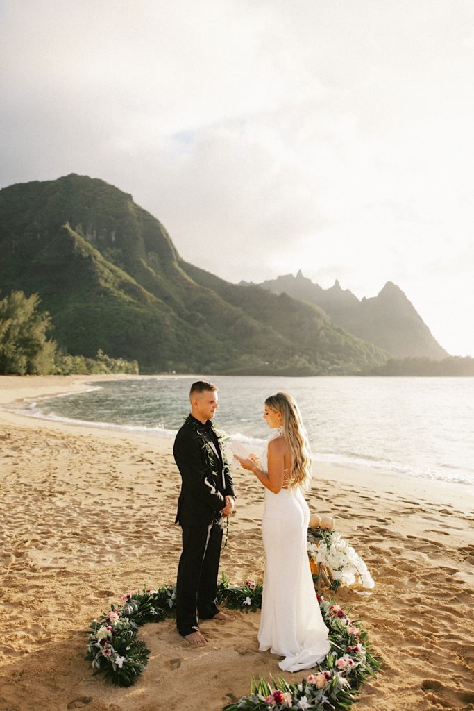 A couple recites their vows to one another on a beach in Hawaii with a circle of flowers on the ground around them.