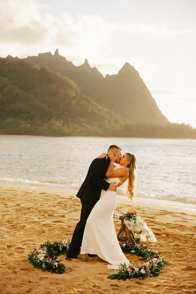 A couple shares their first kiss with one another on a beach in Hawaii with a circle of flowers on the ground around them.