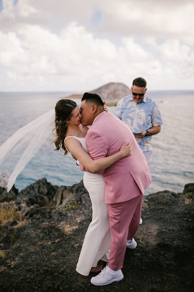 A couple stands kissing, the groom in pink and the bride in a wedding dress with a veil on a cliff in Hawaii overlooking the ocean. 