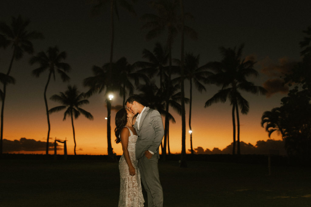A married couple kisses at their wedding in front of a sunset during their reception at Lanikūhonua