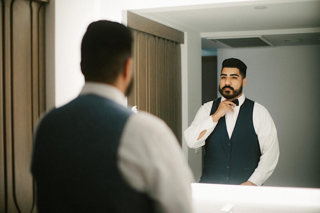 A groom looks in the mirror and adjusts his tie as he gets ready for his wedding day.