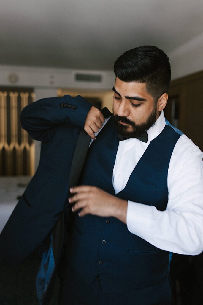 A groom puts on his suit coat in his hotel room as he gets ready for his wedding day.