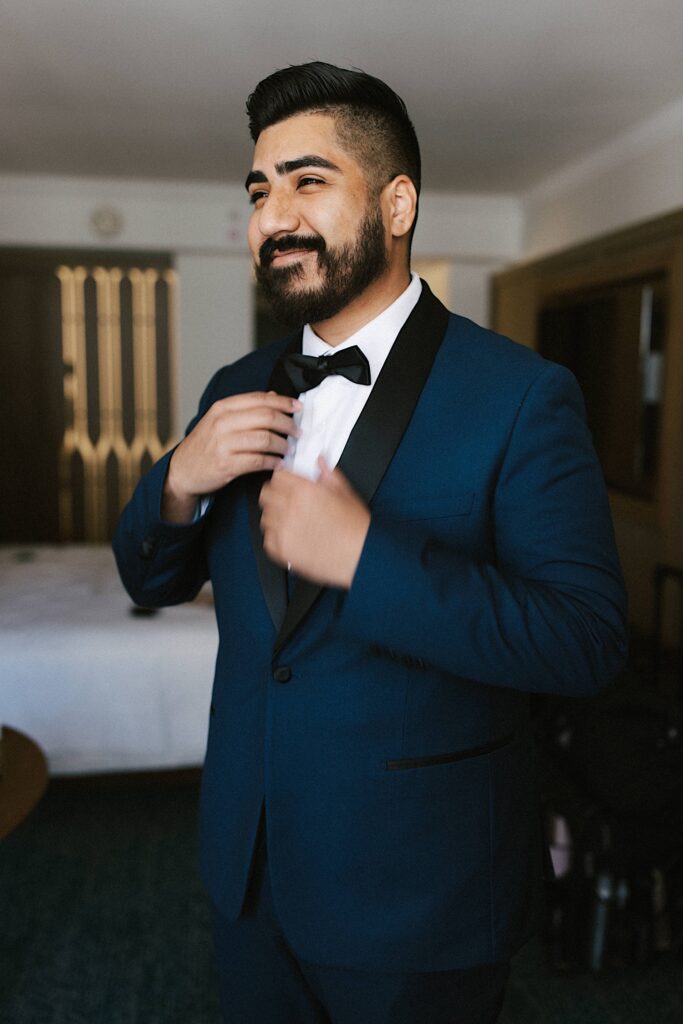 A groom smiles as he adjusts his suit coat in his hotel room while getting ready for his wedding day.