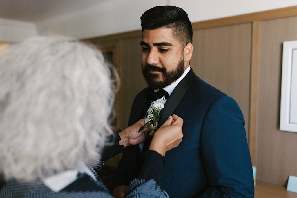A groom stands and smiles as his mother helps pin his carnation to his coat on his wedding day