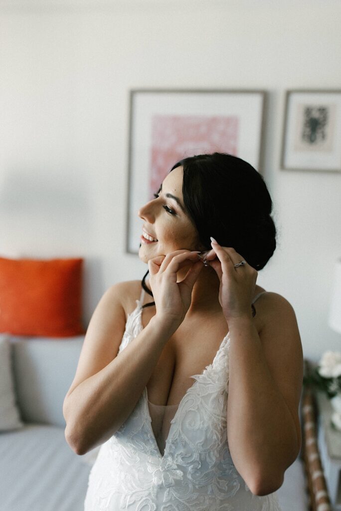 A bride smiles while in her wedding dress as she is putting an earing on.