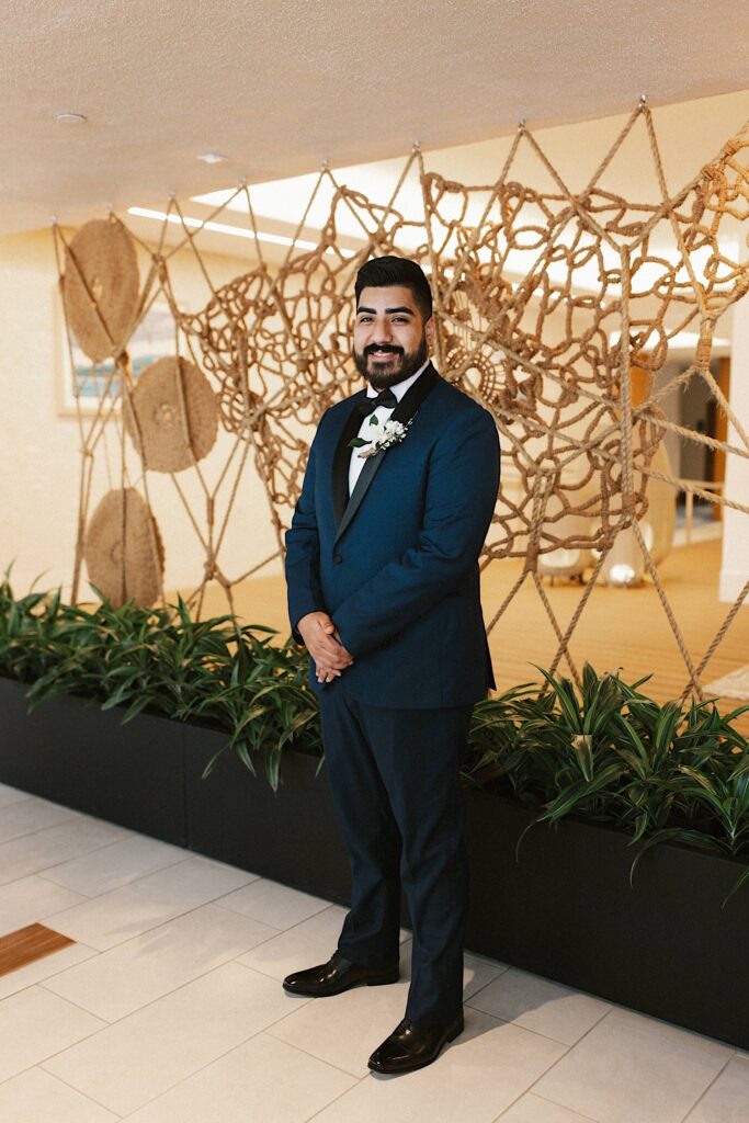 A groom stands with his hands clasped dressed for his wedding day and smiles at the camera