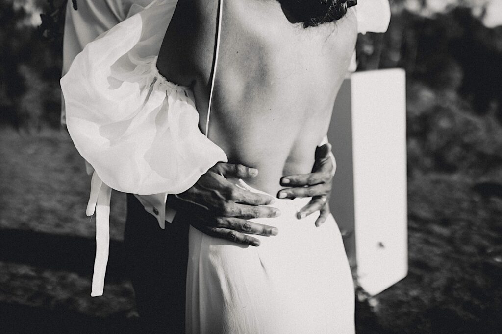 Black and white photo of the back of a bride with the grooms hands around her hips.