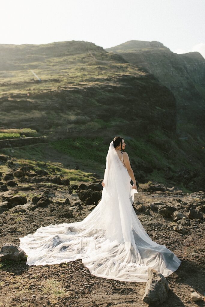 A bride in her wedding dress stands facing away from the camera and looks out over cliffs in Hawaii.