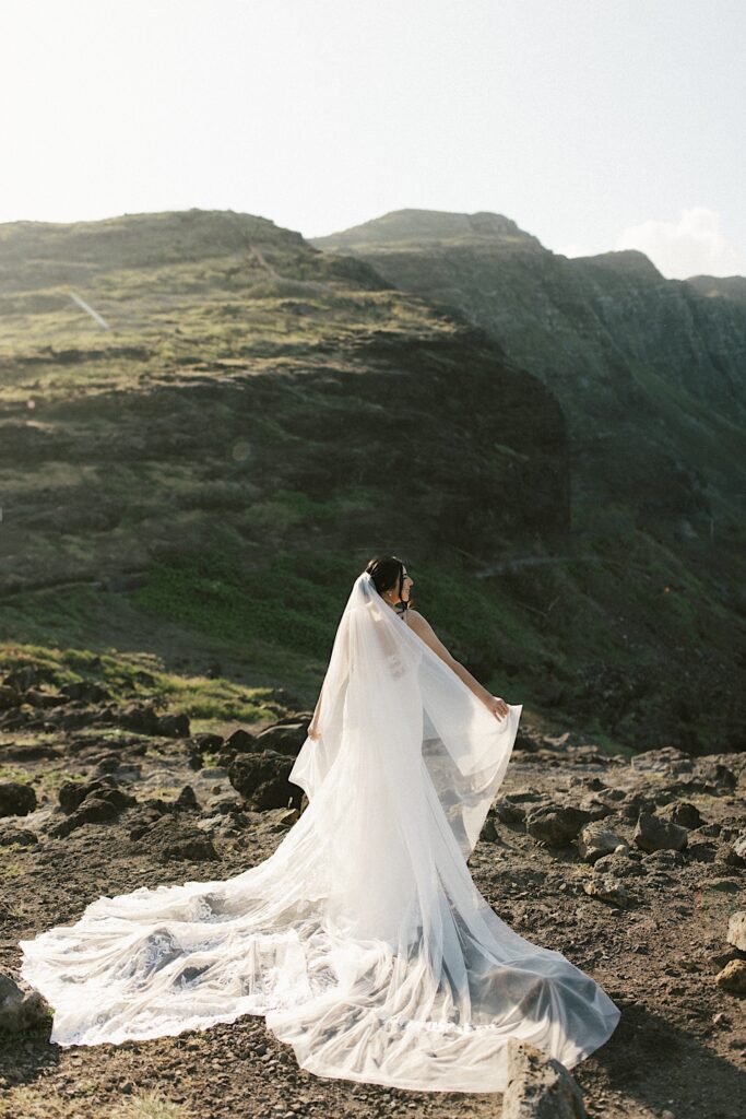 A bride in her wedding dress stands facing away from the camera and looks out over cliffs in Hawaii.