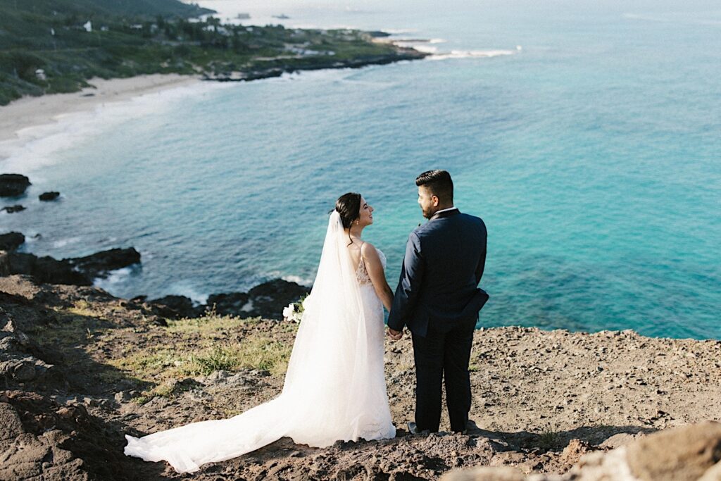 A bride and groom hold hands and smile at one another while on a cliff looking out over Makapuu beach before their elopement.