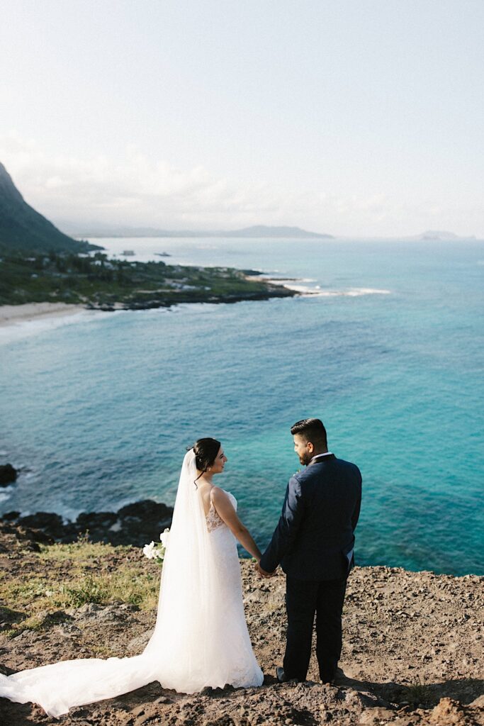 A bride and groom hold hands and smile at one another while on a cliff looking out over Makapuu beach.