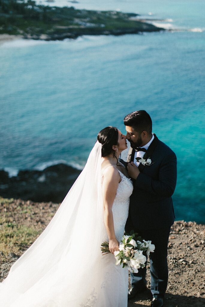 A bride and groom are about to kiss while standing on a cliff looking out over Makapuu beach.