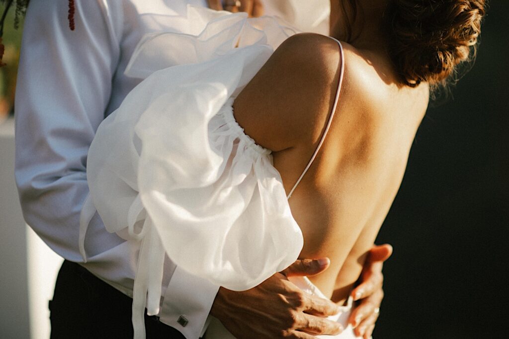 A photo of the back of a bride with the grooms hands around her hips.