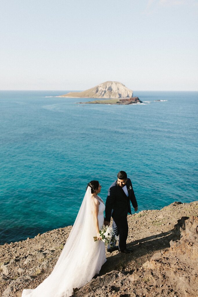 A bride and groom walk hand in hand on a cliff looking out over the ocean in the Hawaiian islands.