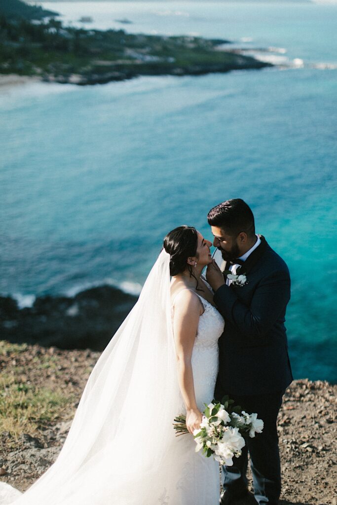 A bride and groom are about to kiss while standing on a cliff looking out over Makapuu beach.